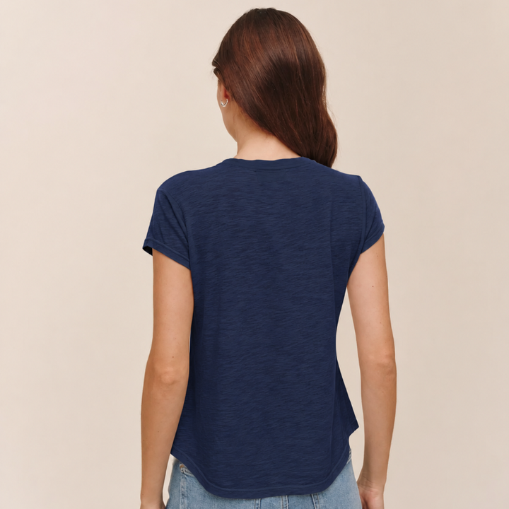 A woman viewed from the back wearing a navy blue short-sleeve t-shirt and light blue jeans, set against a plain beige studio background.