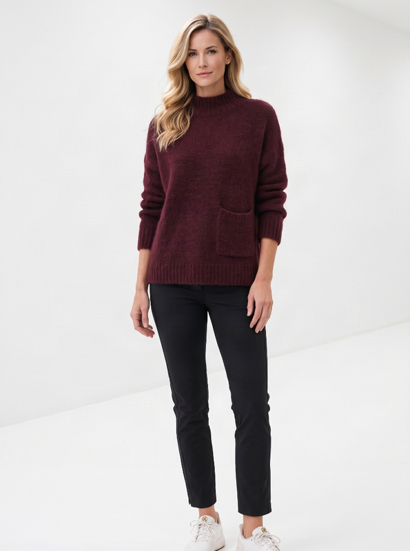 A woman modeling a dark burgundy knit sweater with front patch pockets, paired with black pants and white sneakers, standing in a bright white studio.