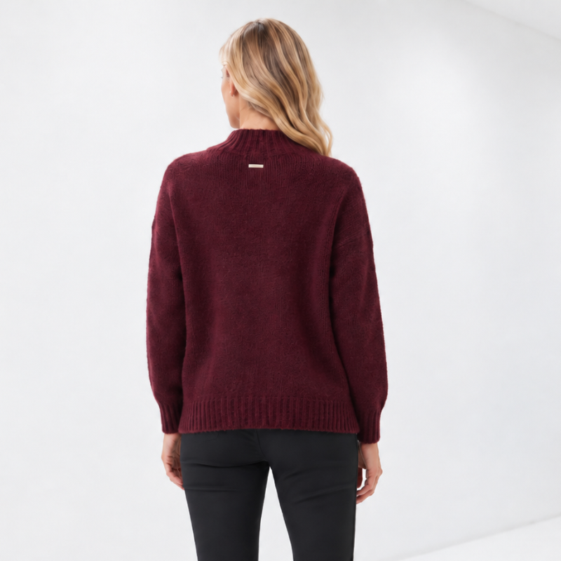 A woman viewed from behind wearing a burgundy knit mock-neck sweater, black slim-fit pants, and white sneakers in a bright white studio setting.