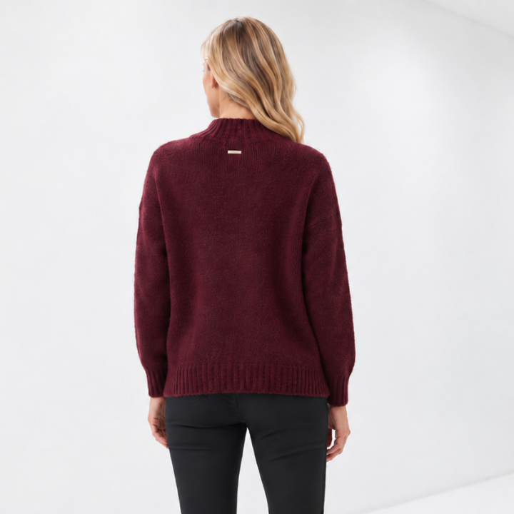 A woman viewed from behind wearing a burgundy knit mock-neck sweater, black slim-fit pants, and white sneakers in a bright white studio setting.