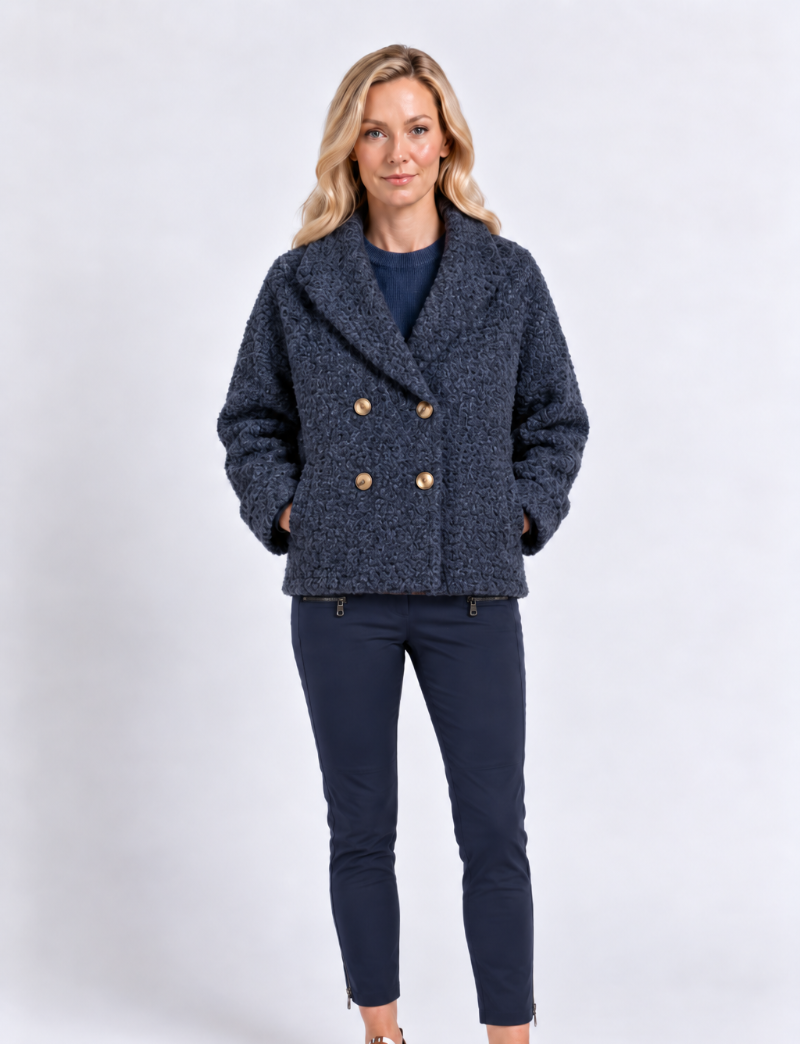 A woman modeling a textured navy blue double-breasted jacket with gold buttons, paired with navy pants and white sneakers, against a light gray studio background.
