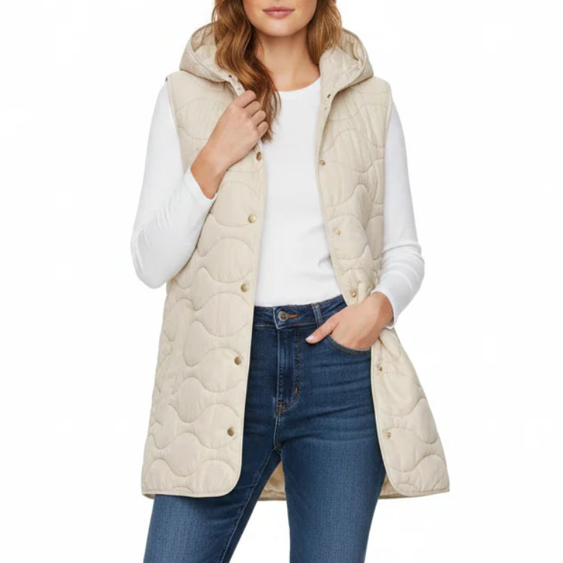 A woman modeling a light beige quilted puffer vest over a white long-sleeve top, paired with blue skinny jeans and white sneakers, against a plain white background.
