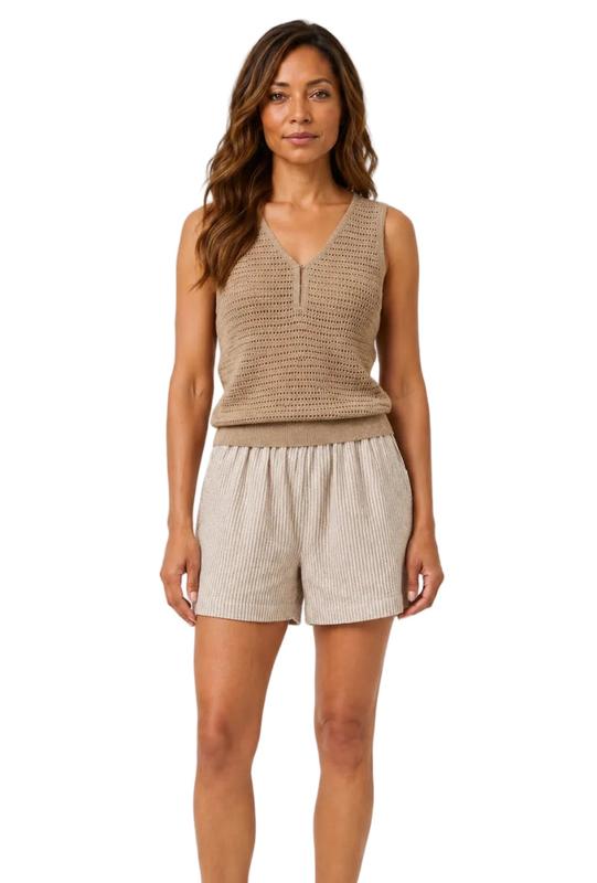 A woman modeling a ribbed knit sleeveless vest in taupe and matching pleated shorts in a light beige, paired with white and green sneakers, against a plain light gray background.